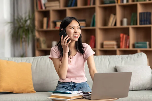 Pozitif Koreli kadın evdeki koltukta otururken telefonla konuşuyor ve dizüstü bilgisayar kullanıyor.