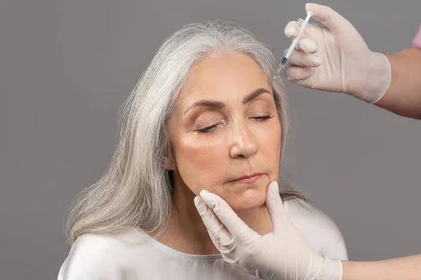 Charming senior woman getting beauty injection, receiving hyaluronic acid or collagen shot on grey studio background — Foto de Stock