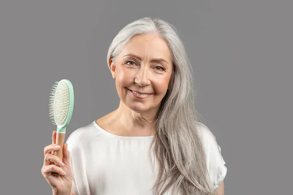 Aging gracefully. Charming senior lady with silky long hair holding brush and smiling on grey studio background — Fotografia de Stock