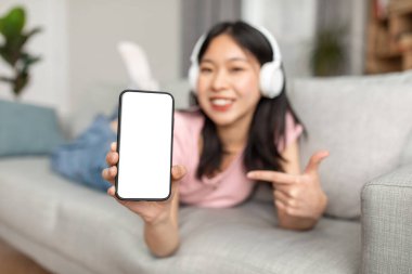 Great musical app. Happy asian lady showing phone empty screen, wearing wireless headphones, lying on sofa at home
