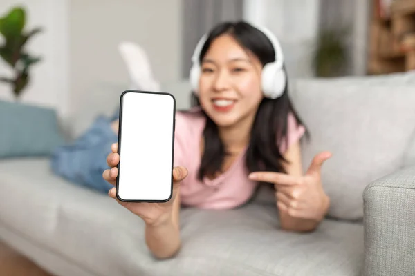 Great musical app. Happy asian lady showing phone empty screen, wearing wireless headphones, lying on sofa at home — ストック写真