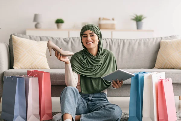 Mladá Arabka v hidžábu drží digitální tablet a novou botu, sedí na podlaze mezi dárkovými taškami, psaní recenze — Stock fotografie