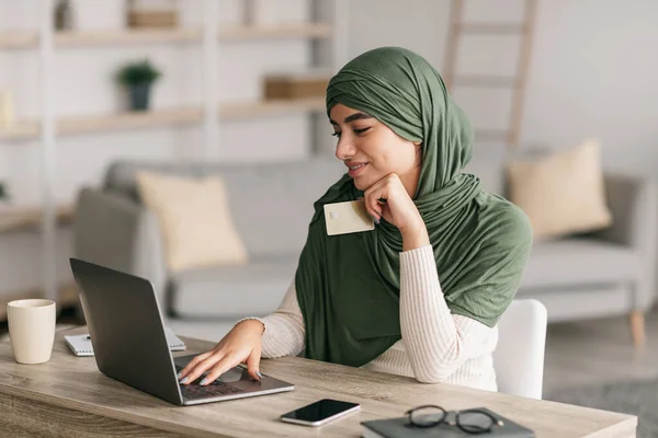不用现金支付。阿拉伯妇女头戴头巾，手持信用卡，使用手提电脑，从家里上网购物 — 图库照片