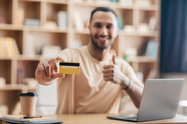 Vzrušený arabský muž, který provádí online platby pomocí notebooku a kreditní karty, ukazuje karty do kamery a ukazuje palec nahoru — Stock fotografie