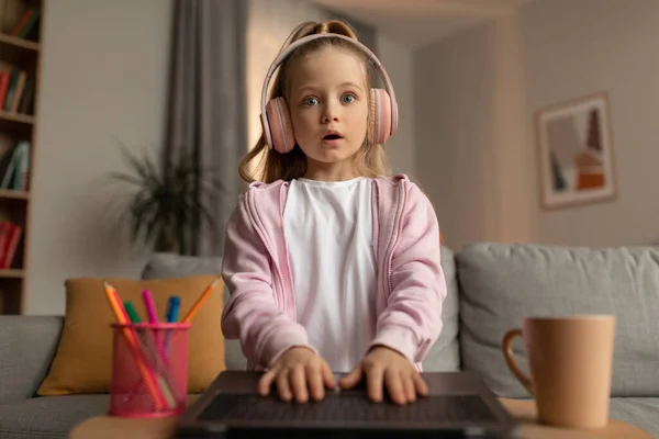Shocked Little Schoolgirl Looking At Laptop Wearing Headphones At Home — Fotografia de Stock