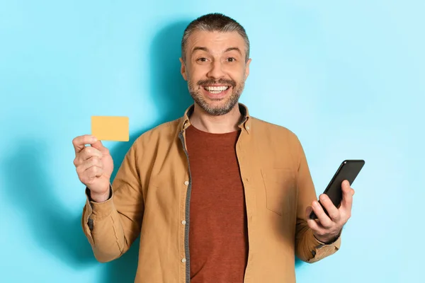 Middle Aged Man Holding Cellphone Showing Credit Card, Blue Background — Stockfoto