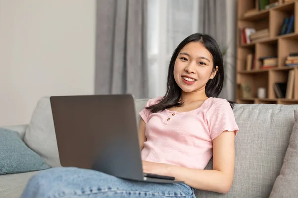Portrait of happy asian lady sitting on sofa with laptop on laps and smiling at camera, having distance job — ストック写真