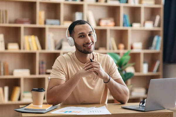 Portrait of arab man in wireless headset sitting at desk with laptop, looking and smiling at camera, working at home —  Fotos de Stock