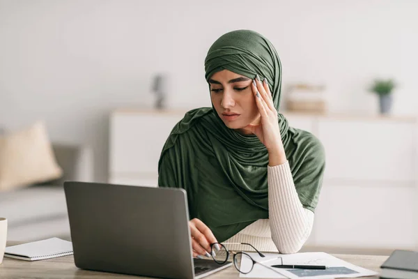 Unhappy young Arab female freelancer in hijab feeling tired of working on laptop, suffering from headache at home office — Stok fotoğraf