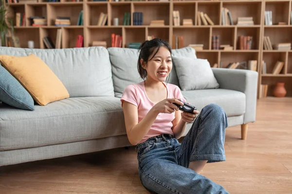 Happy asian lady holding joystick and playing videogames while resting on sofa at home, free space — Foto de Stock