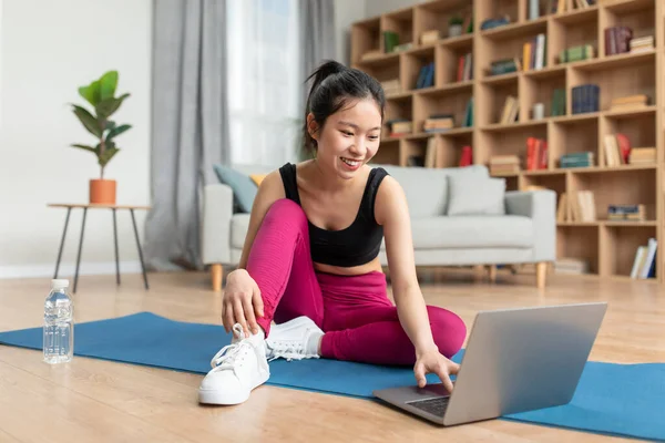 Happy korean fitness blogger looking at laptop, chatting and choosing online workout lesson at home, sitting on yoga mat — Stockfoto