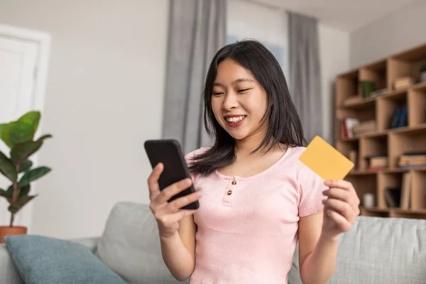 Fröhliche koreanische Dame mit Handy und Kreditkarte, die Waren oder Dienstleistungen im Internet kauft, sitzt zu Hause auf dem Sofa — Stockfoto