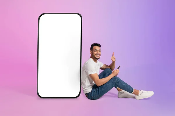 Jovem árabe cara sentado perto de grande celular com tela branca em branco, usando dispositivo móvel, mostrando polegar acima gesto, mockup — Fotografia de Stock