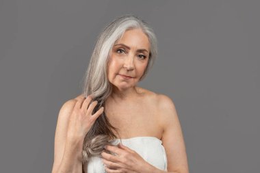 Feminine senior lady with perfect smooth skin wearing towel after bath, posing to camera over grey studio background