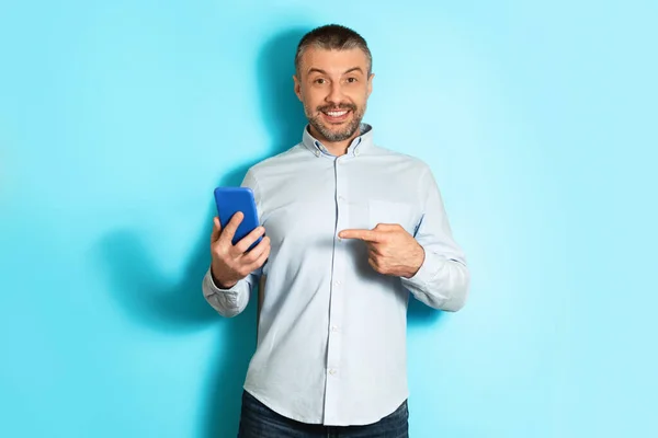 Middle Aged Male Pointing At Phone Advertising App, Blue Background — Stok Foto