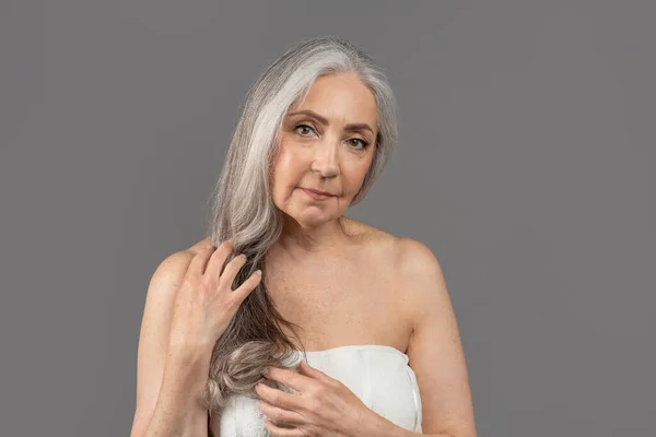 Senhora sênior feminina com pele lisa perfeita vestindo toalha após o banho, posando para câmera sobre fundo estúdio cinza — Fotografia de Stock