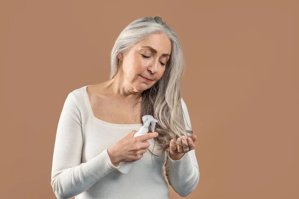 Szép idős nő alkalmazó hajápoló spray vagy természetes illóolaj gyönyörű szürke haj felett barna stúdió háttér — Stock Fotó