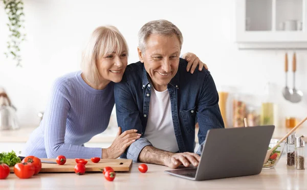 Loving senior couple cooking together at home, using laptop — Stockfoto