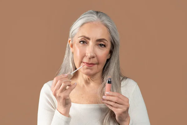 Beauty portrait of senior woman applying lip gloss and looking at camera on brown studio background. Organic makeup — Stock Fotó