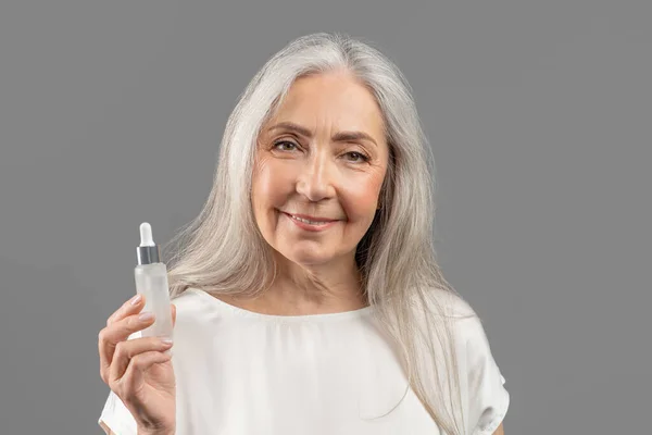 Szépségápolási eljárás. Bájos érett nő bemutató üveg anti-aging szérum szürke stúdió háttér — Stock Fotó