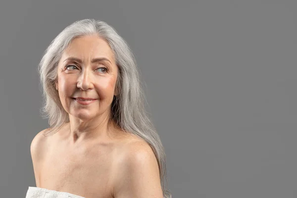 Portrait of charming senior lady with smooth silky skin looking aside at empty space, wearing towel on grey background — 스톡 사진