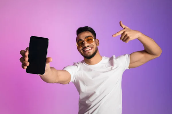Leende ung arabisk man i solglasögon pekar på smartphone med tom skärm i neonljus, selektivt fokus. Uppställning — Stockfoto