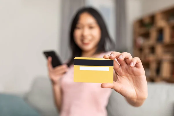 Asijská dáma ukazuje kreditní kartu na kameru, držení smartphone a nakupování v internetovém obchodě z domova, selektivní zaměření — Stock fotografie