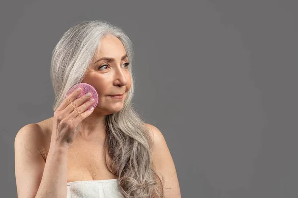 Mulher sênior encantadora com cabelo longo bonito segurando esponja cosmética perto de seu rosto no fundo do estúdio cinza, espaço de cópia — Fotografia de Stock