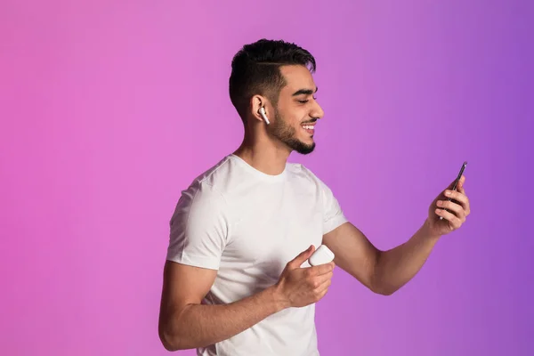 Happy young Arab guy with smartphone listening to music in wireless earphopnes, using new mobile app in neon light — Stok fotoğraf