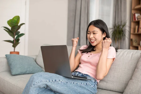 Excited korean lady sitting on couch with laptop, lifting hands up, gesturing YES and celebrating success at home — Foto Stock
