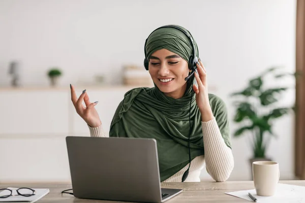 Pozitív fiatal arab nő hidzsáb és fejhallgató video chat, amelynek online konferencia laptop otthon — Stock Fotó