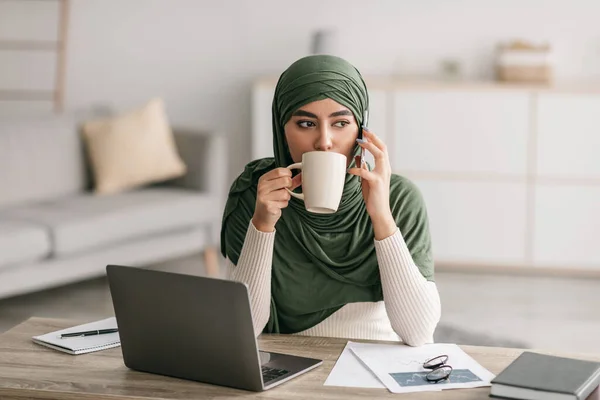 Fiatal arab nő hidzsáb dolgozik online otthoni irodában, laptoppal, hívja az okostelefon és kávézás — Stock Fotó