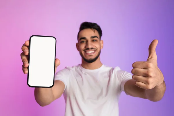 Happy yougn Arab guy holding cellphone with empty white screen and showing thumb up gesture in neon light, mockup — Fotografia de Stock