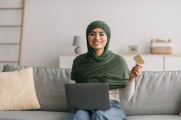 Happy young Arab woman using laptop and credit card to purchase things on internet, buying products online from home