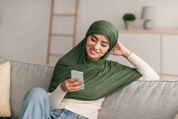 Positive young Arab lady in hijab sitting on couch, having video call with friends or lover, using modern smartphone at home — Stockfoto