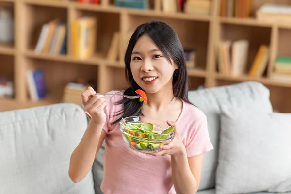 Healthy nutrition concept. Young asian lady eating yummy vegetable salad on sofa at home, smiling at camera — Stockfoto