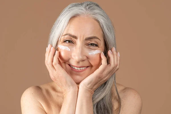Mature beauty. Charming senior woman posing with face cream under her eyes, smiling at camera over brown background — Stok fotoğraf