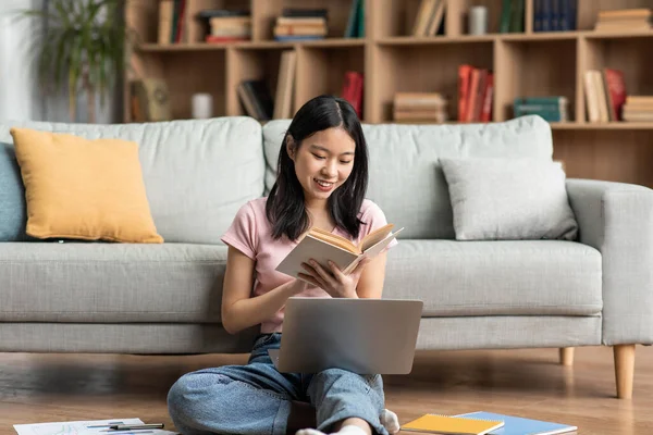 Happy asian lady studying online from home, reading book and using laptop at home, sitting on floor near sofa — Foto Stock