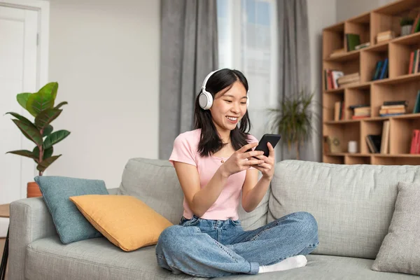 ソーシャルネットワークでのオンラインチャット。幸せな若い韓国人女性のヘッドフォンで入力するスマートフォン、ソファの上に休んで — ストック写真