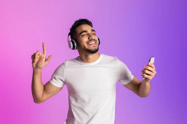 Hombre árabe joven guapo escuchando música en los auriculares, utilizando la nueva aplicación de teléfono inteligente y bailando en luz de neón —  Fotos de Stock