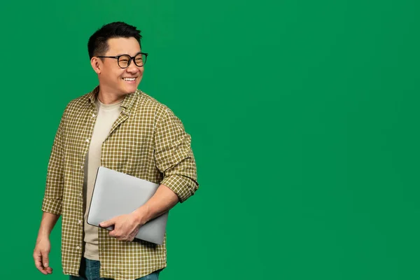 Онлайн-пропозиція. Зрілий азіатський чоловік тримає ноутбук і озирається назад на вільний простір над зеленим студійним фоном — стокове фото