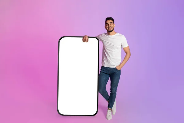 Young Arab man leaning on big cellphone with empty screen, offering space for your ad, mobile app or website, mockup —  Fotos de Stock