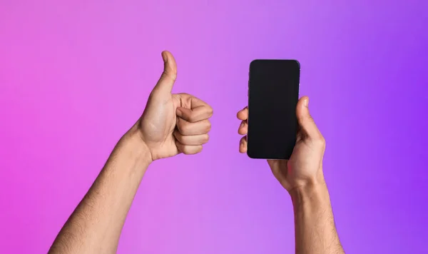 Обрезанный вид молодого парня, показывающего смартфон с пустым экраном и жестикулирующего большим пальцем вверх, рекомендующий макет мобильного приложения — стоковое фото