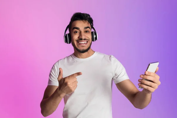 Porträtt av funky leende ung arabisk man i trådlösa hörlurar pekar på smartphone i neon ljus — Stockfoto