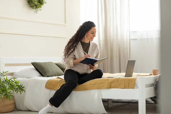 Leende kvinna sittande på sängen, med dator skriva i anteckningsbok — Stockfoto