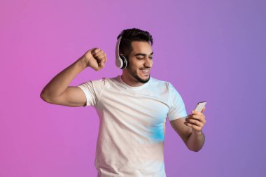 Mutlu genç Arap adam kulaklıkla müzik eşliğinde dans ediyor, akıllı telefon ekranına bakıyor, neon ışıkta piyango kazanıyor.