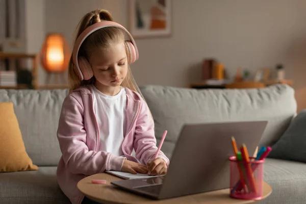 Schoolgirl Learning Using Laptop Taking Notes Wearing Earphones At Home — Fotografia de Stock