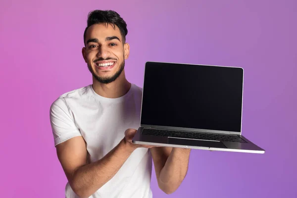 Smiling millennial Arab man showing laptop computer with empty black screen in neon light, mockup for website or ad — Foto de Stock