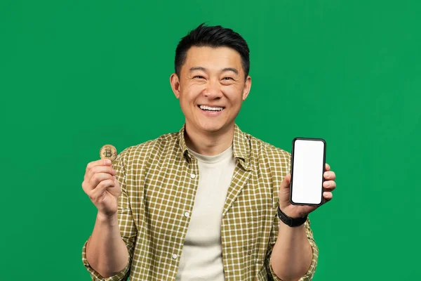 Mature asian man holding golden bitcoin coin and showing cellphone with white screen, recommending online trading app — Fotografia de Stock