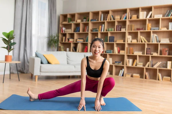 Ev antrenmanı. Genç Asyalı bayan oturma odasında yoga minderinde bacaklarını açıyor ve kameraya gülümsüyor.
